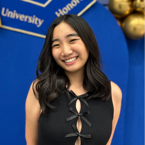 A woman woman with long black hair in a black top, posing in front of a blue backdrop with text that reads "University Honors".
