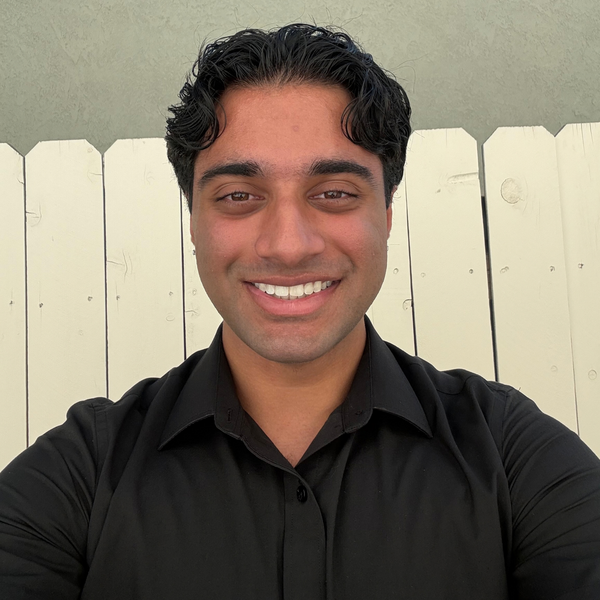 Portrait of a man with short dark hair wearing a dark button-up shirt, facing the camera with a white wooden fence in the background.