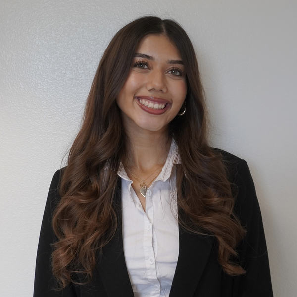 Smiling woman with long wavy hair, wearing a white button top and black blazer posing in front of a light background.