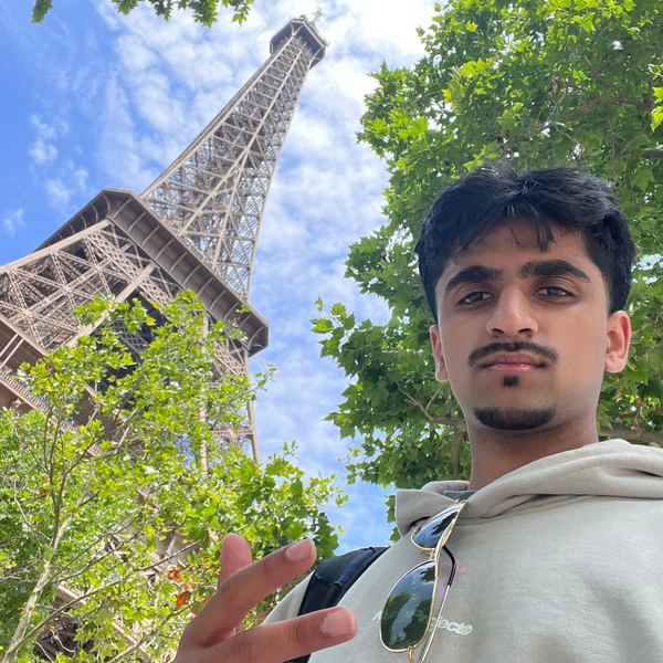 A photo of a man with dark hair posing outdoors surrounded by green foliage and the Eifel Tower.