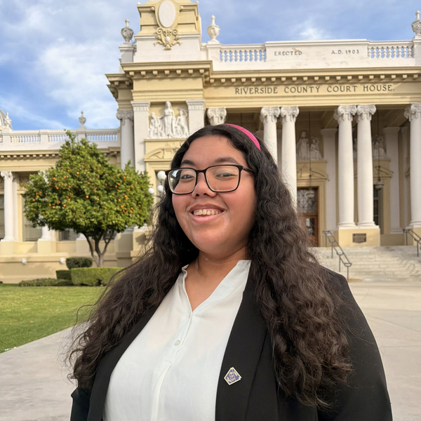 Woman outdoors in front of a tall stone building with columns, standing on a paved walkway and smiling toward the camera in a white shirt and black blazer.